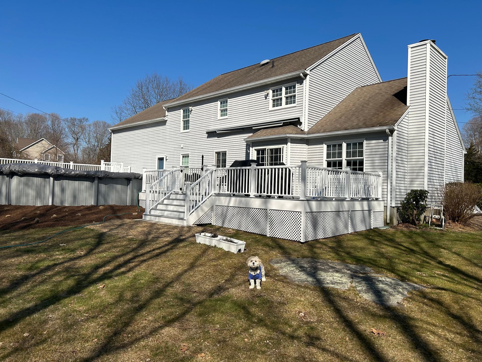 Backyard with two-story white house, wooden deck, and small dog wearing a sweater on a sunny day.