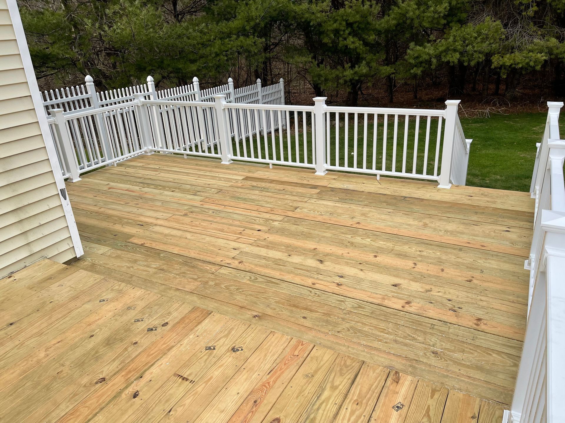 Wooden deck with white railing, attached to a house. Green trees and grass in the background.
