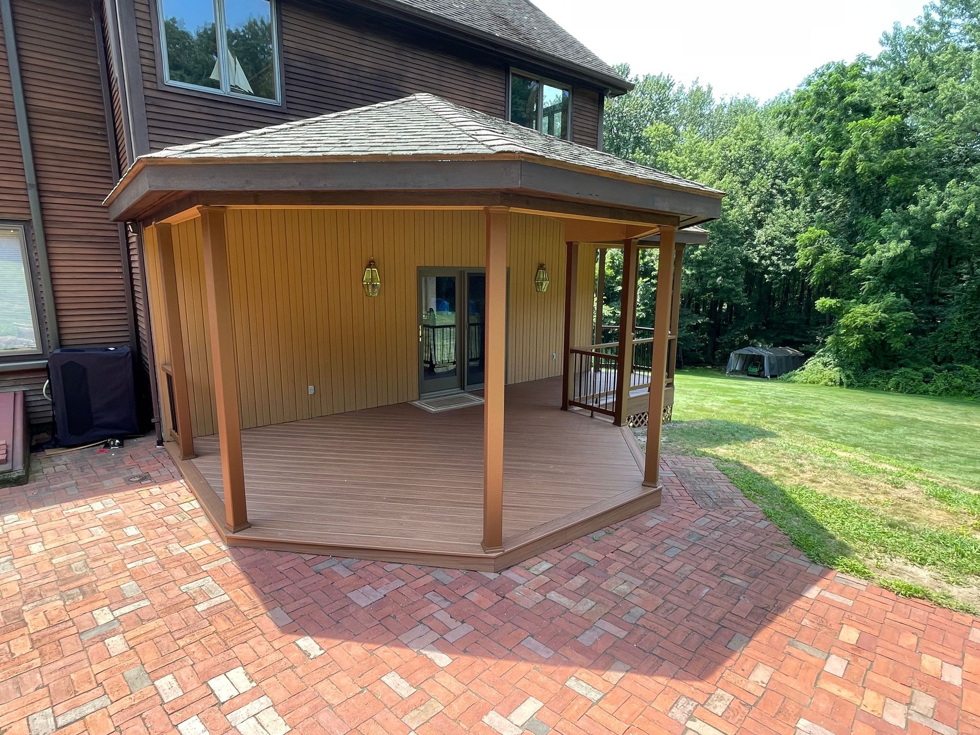 Wooden patio attached to a two-story brown house with red brick pathway and green yard.