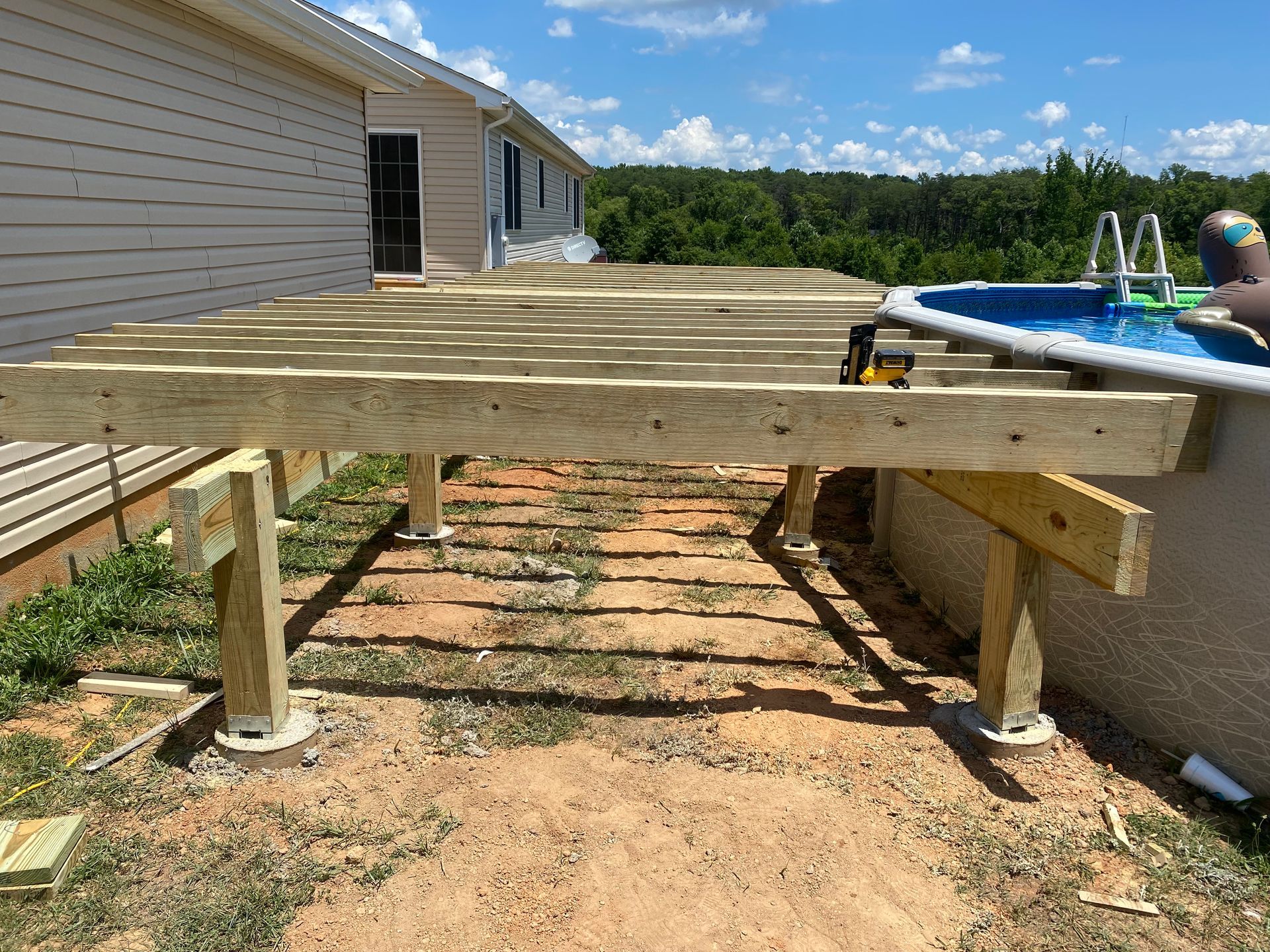Deck construction: wooden frame built around an above-ground pool, near a house on a sunny day.