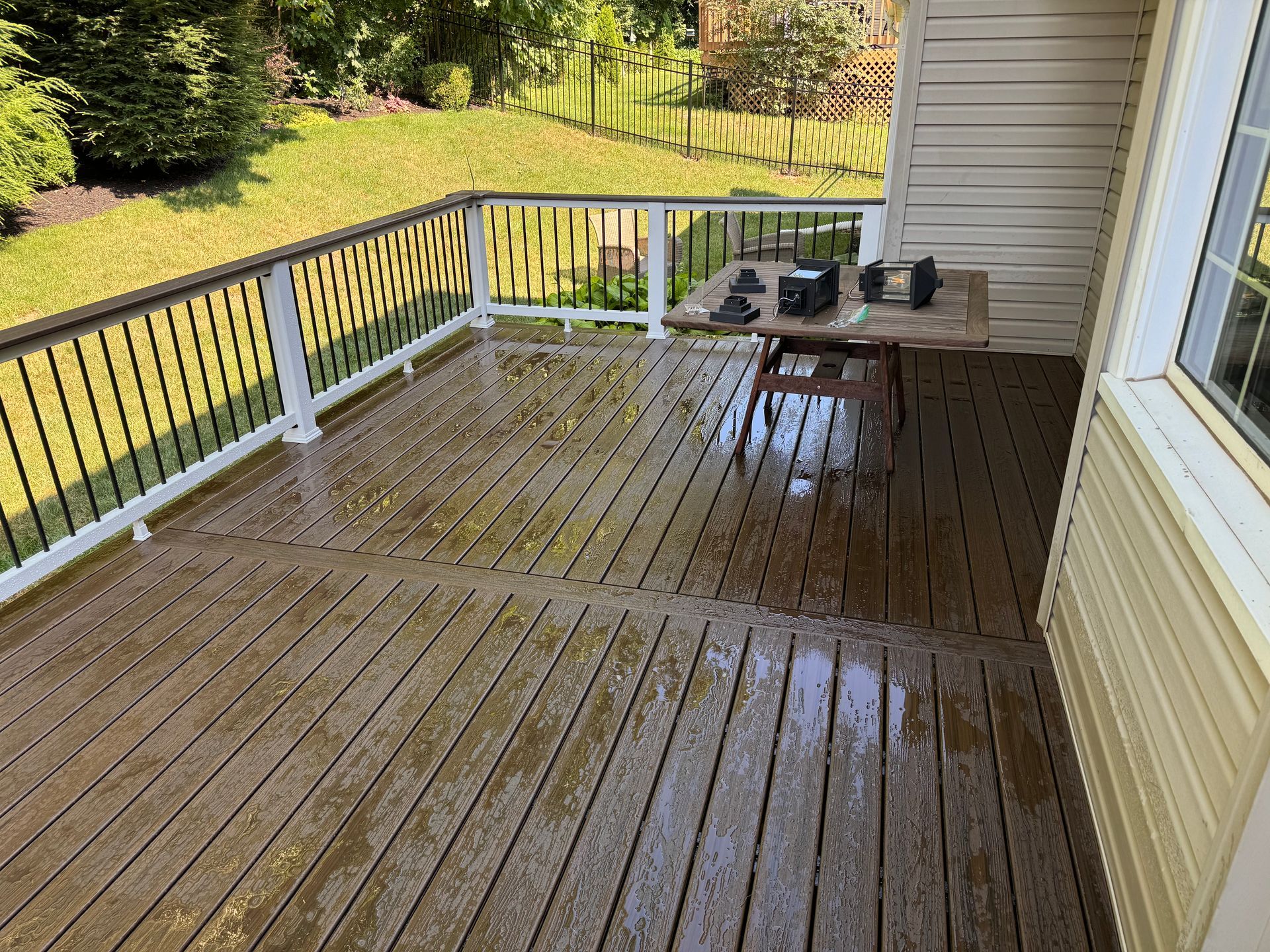 Wooden deck with wet planks and a table, surrounded by railing and grassy yard.
