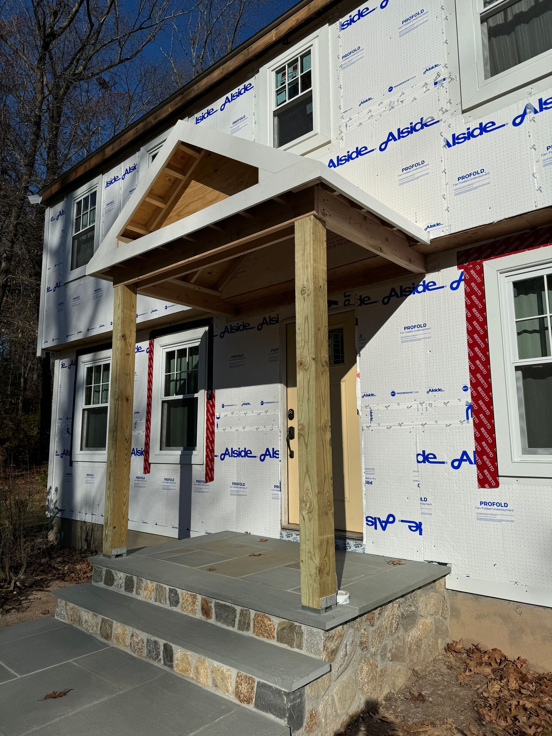 Two-story house under construction with a porch. Blue stone steps lead to the front door under a wood-framed canopy.