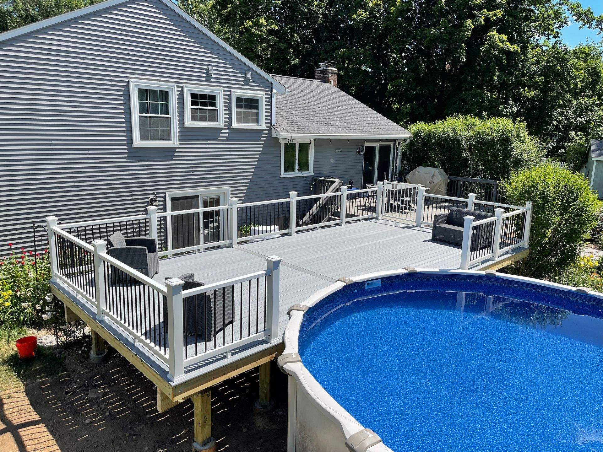 Gray house with a deck and above-ground pool. Black furniture on deck with white railing. Blue water in pool.