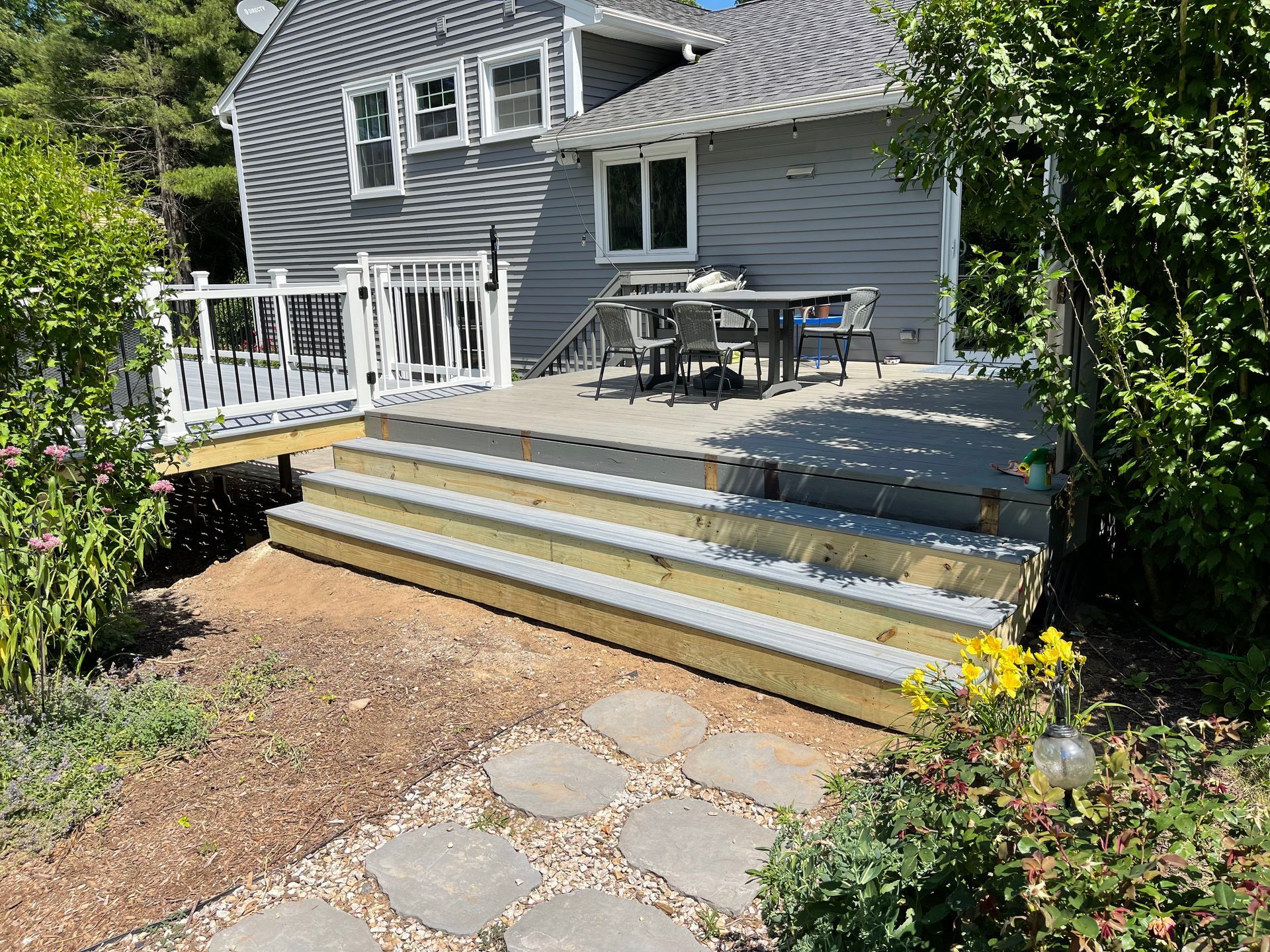 A gray house with a wooden deck and steps, a patio table and chairs, and a stone pathway.