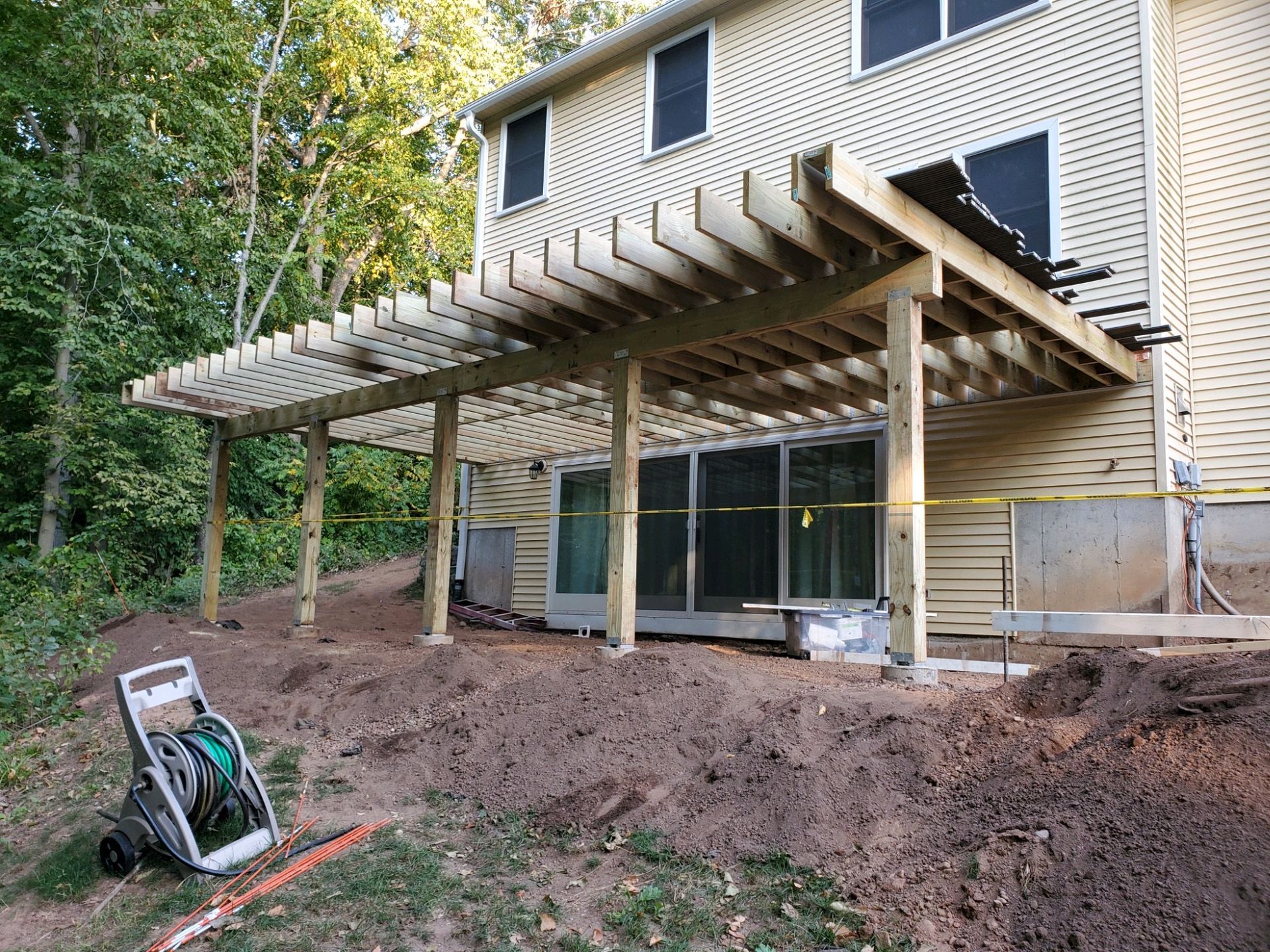 A partially built wooden deck with a pergola, next to a two-story house with a sliding glass door.