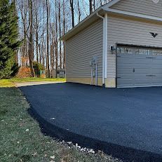A newly paved black asphalt driveway leads to a garage with beige siding, adjacent to a grassy lawn and a line of trees.