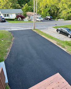 An elevated view of a freshly paved black asphalt driveway leading toward a quiet road with houses and parked cars.