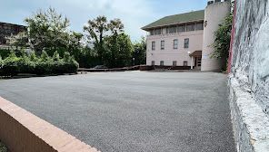 A paved courtyard in front of a light-colored, two-story building with a tower on the right and a stone wall on the left.