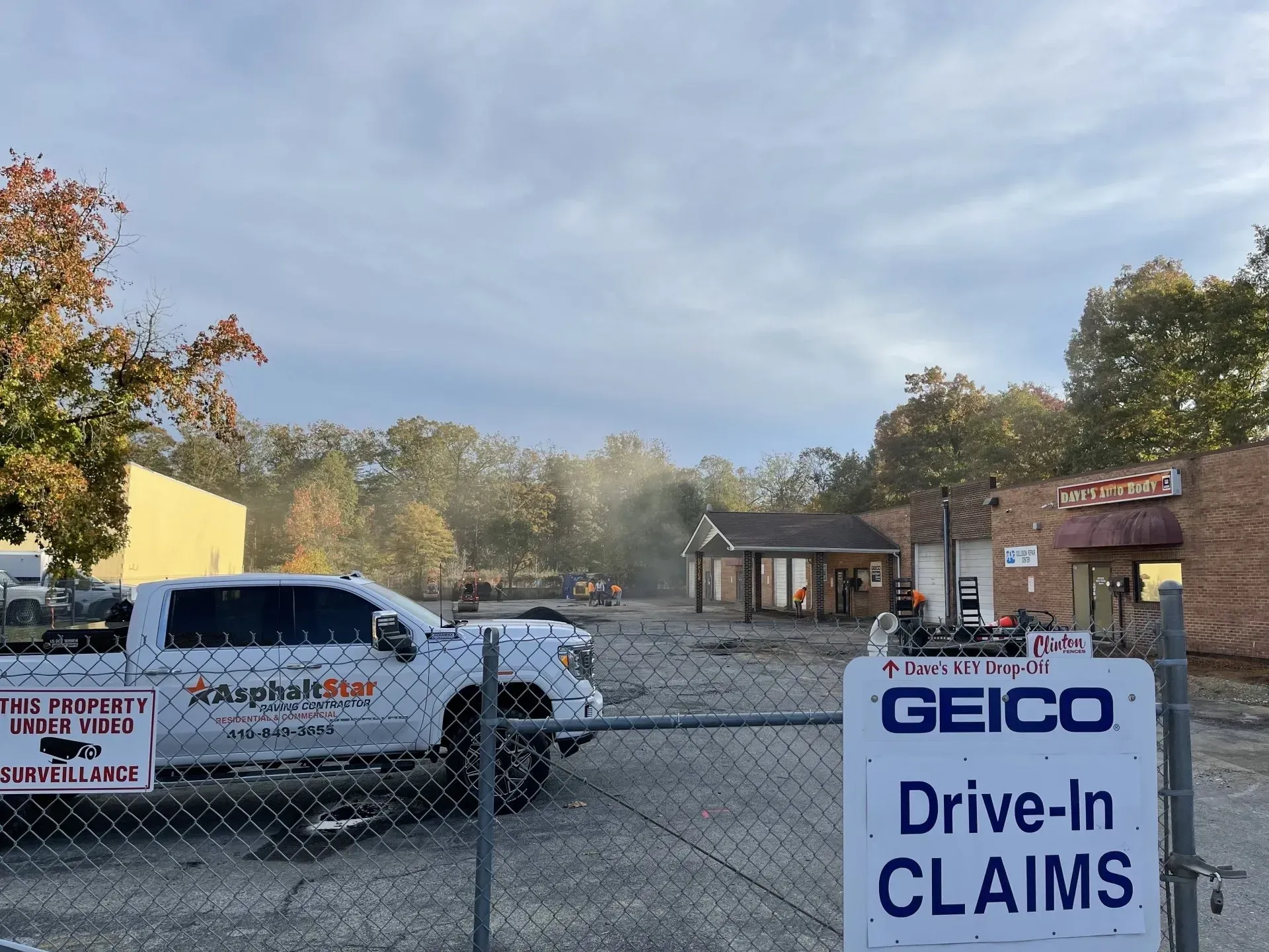 A white truck parked in a gravel lot next to a small commercial building behind a fence with a GEICO Drive-In Claims sign.