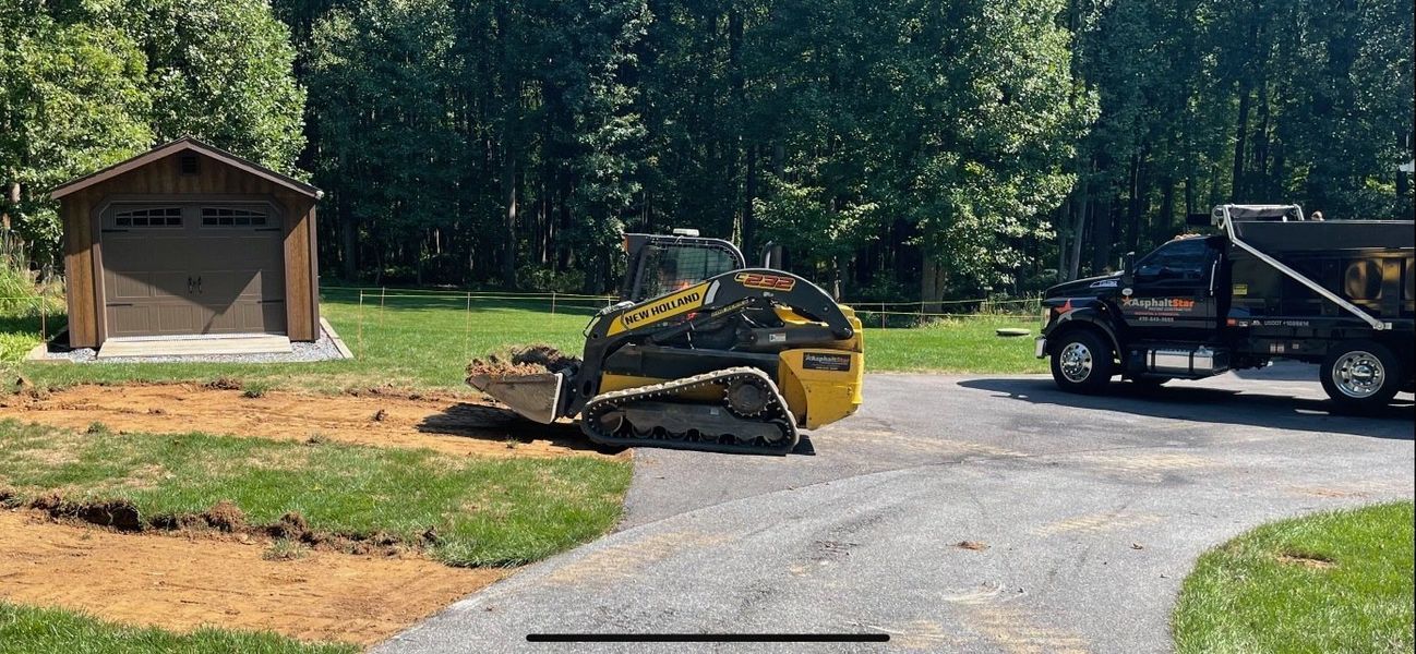 A yellow skid-steer loader scoops dirt next to a gravel driveway, with a dark dump truck and shed in the background.