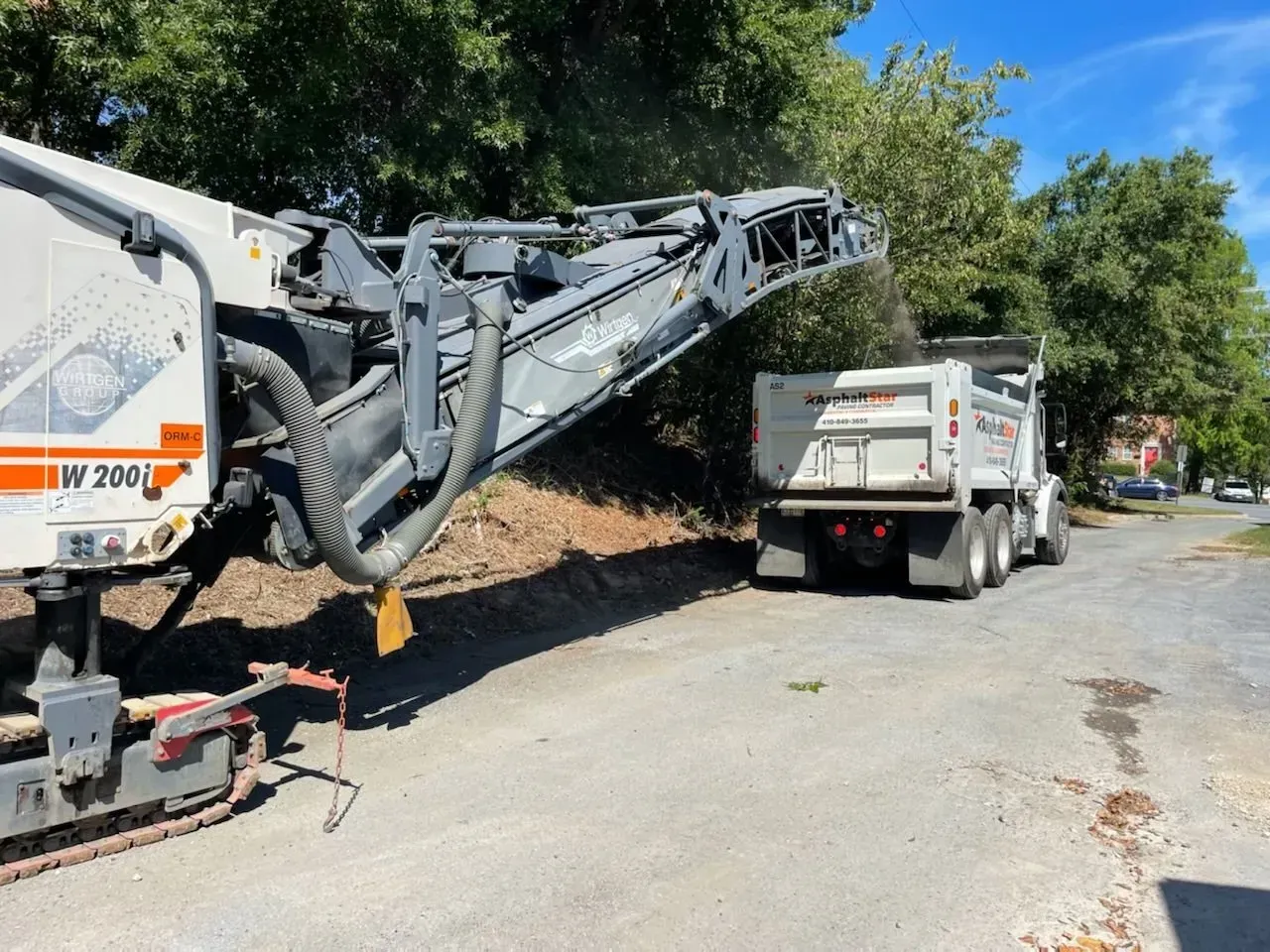 A Wirtgen W 200 road milling machine loads asphalt debris into a white dump truck on a street under trees.