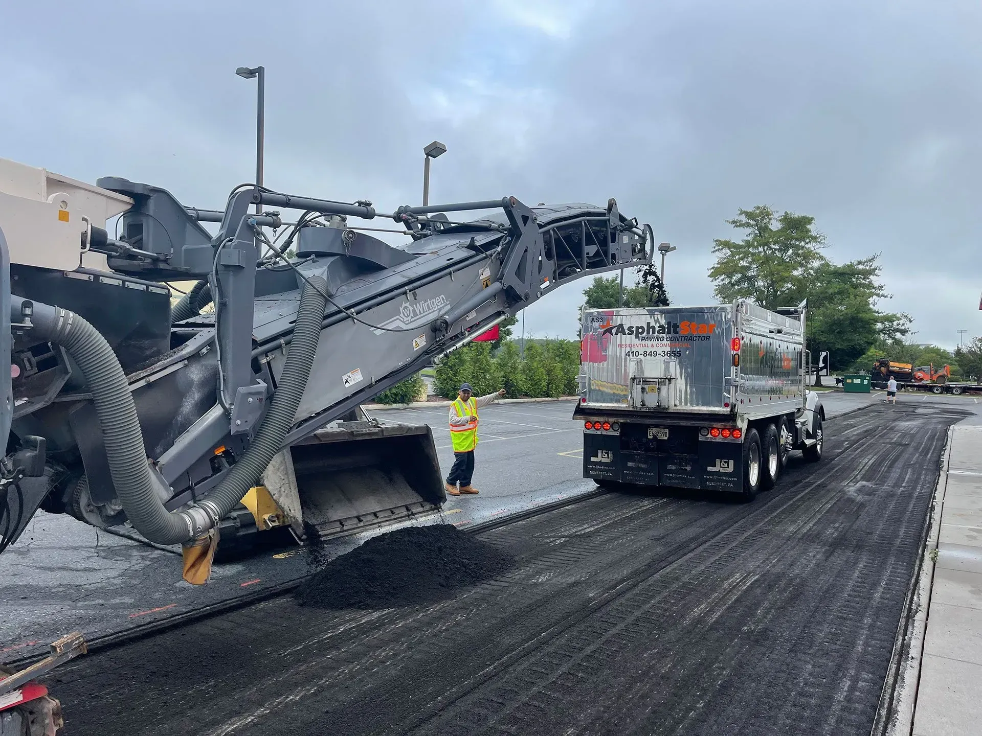 Construction equipment mills old asphalt, loading the debris into a dump truck on a grey, overcast day.