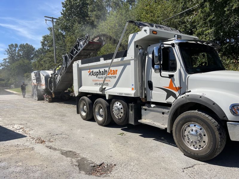 An AsphaltStar dump truck sits on an asphalt road as a milling machine loads it with removed pavement material.