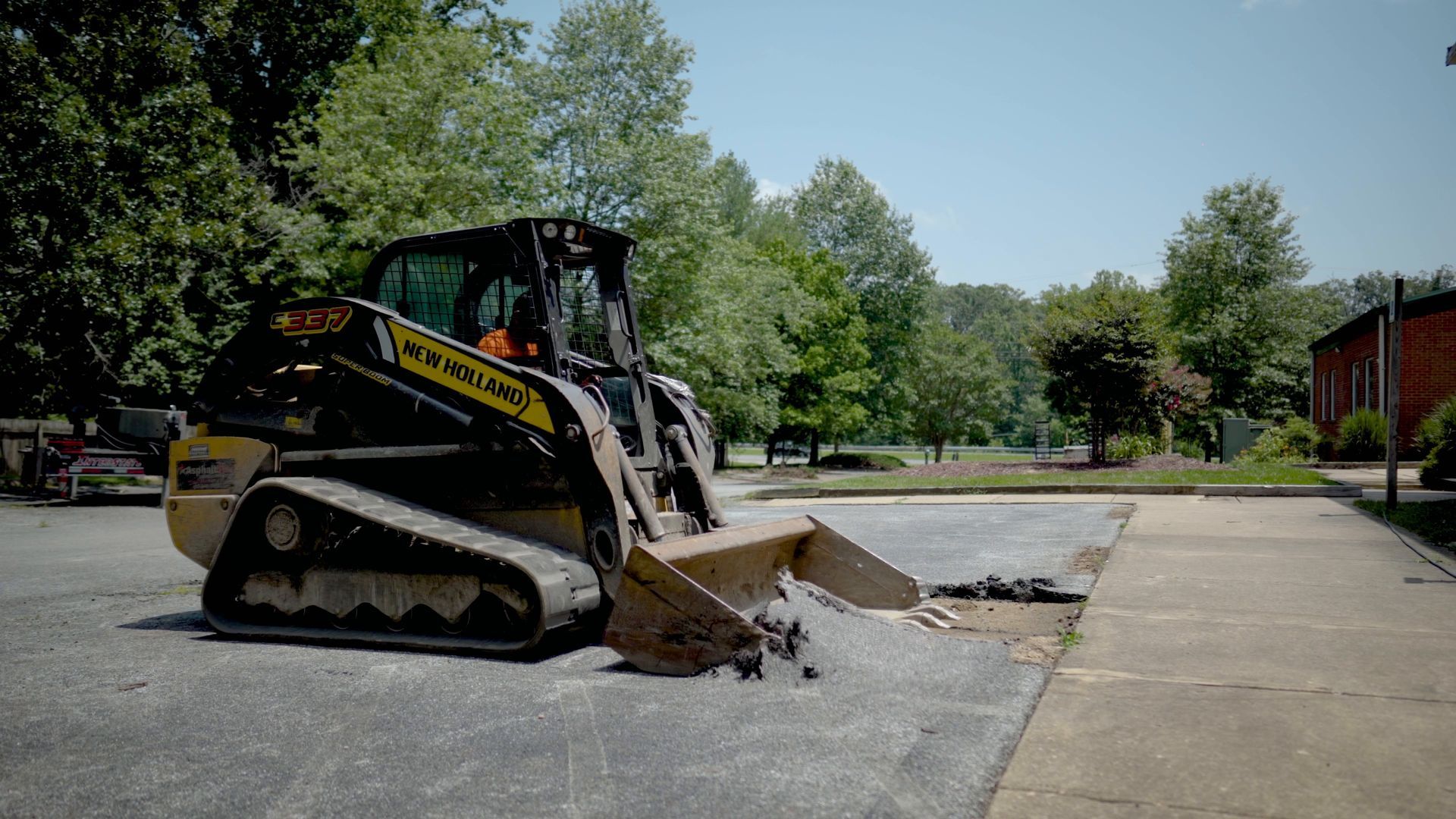 A yellow New Holland skid-steer loader with tracks parked on an asphalt surface, preparing to clear a pile of rubble.