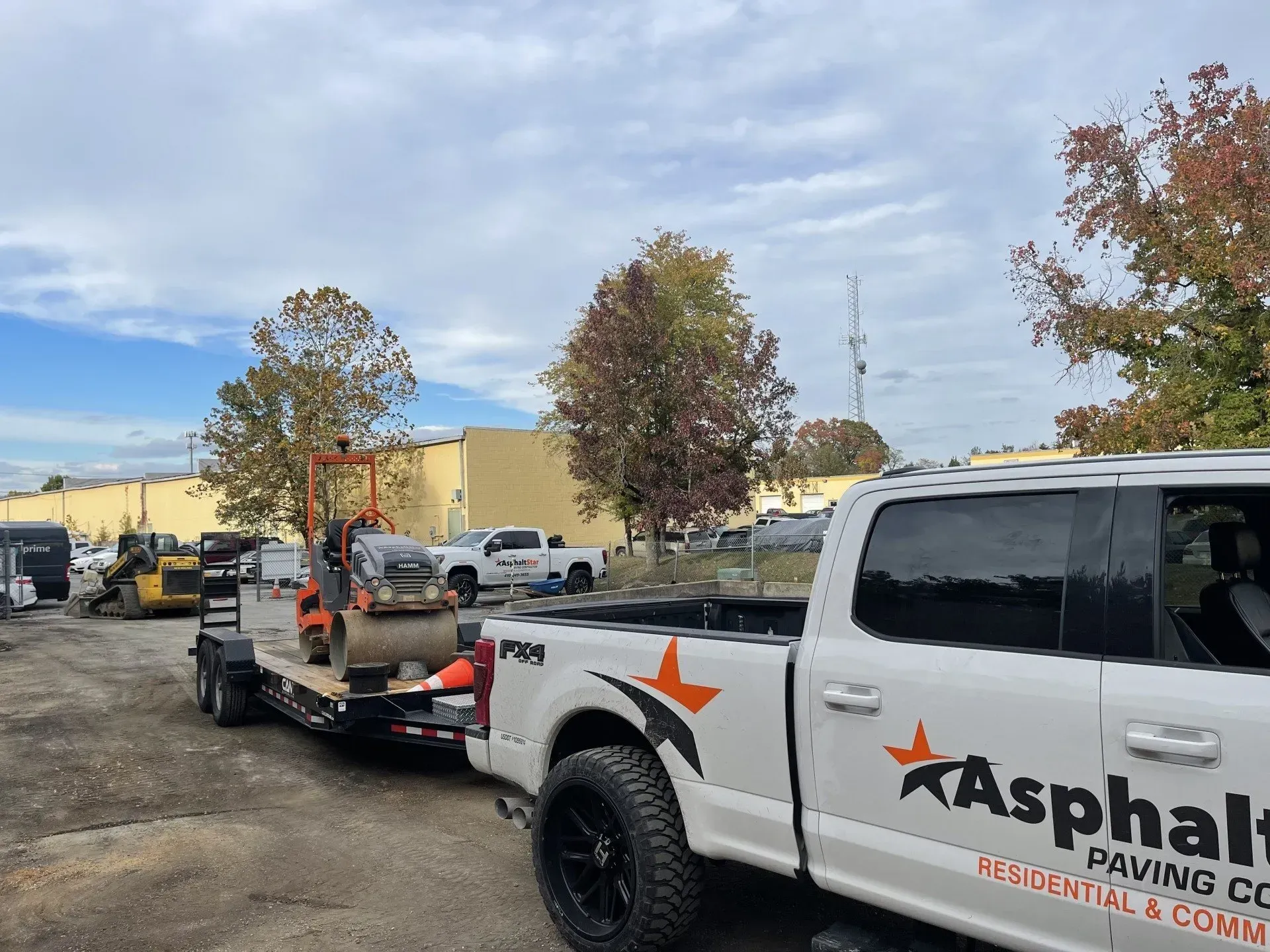 A white Asphalt Paving Co. pickup truck towing a trailer with a construction roller on a gravel lot near a large building.