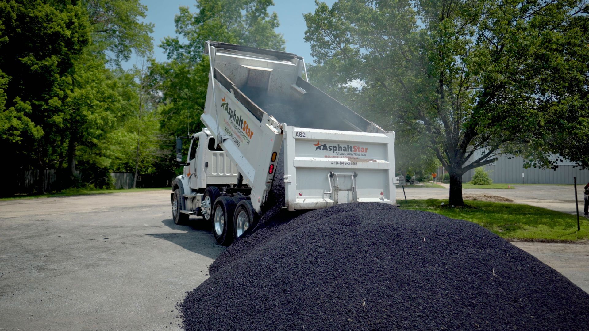 A white dump truck parked in a gravel lot, its raised bed dumping a large pile of black rubber mulch.