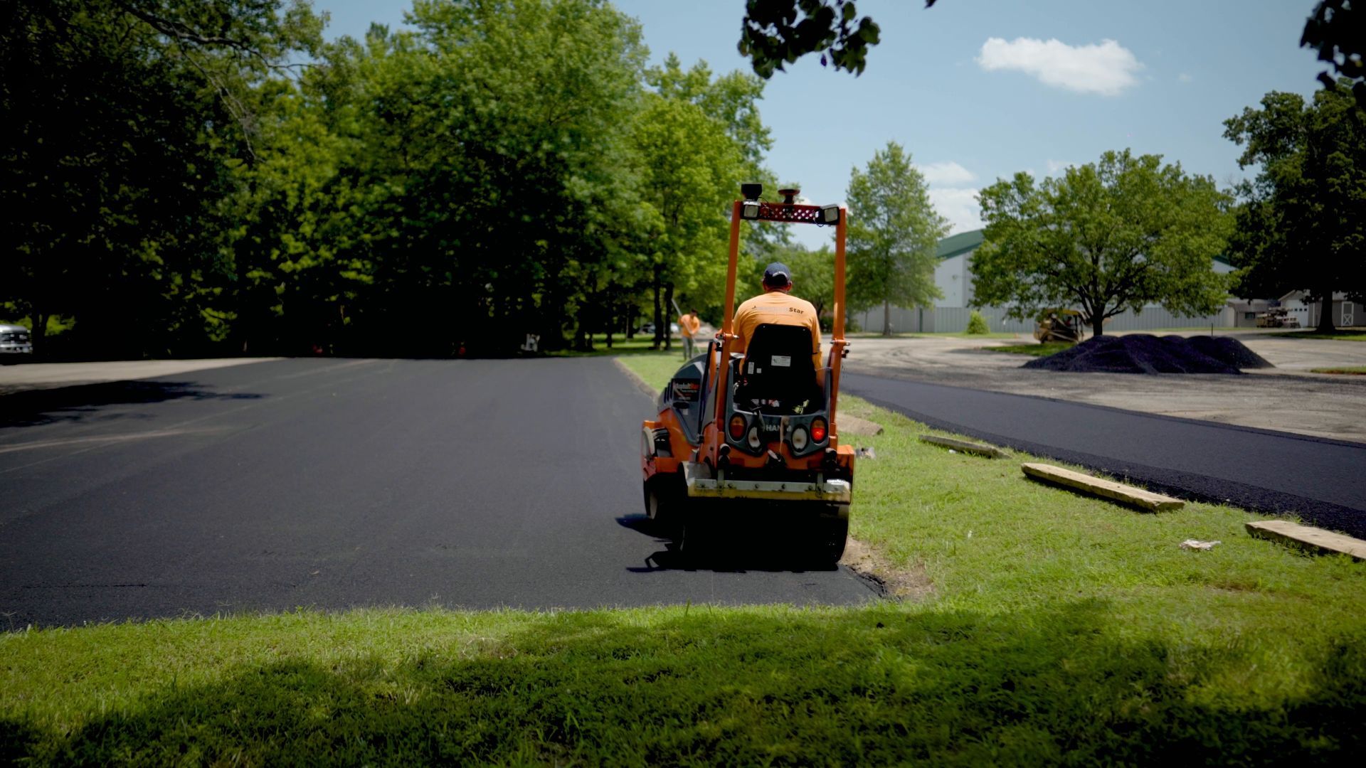 An orange pavement roller operated by a worker is smoothing a freshly paved black asphalt parking lot.