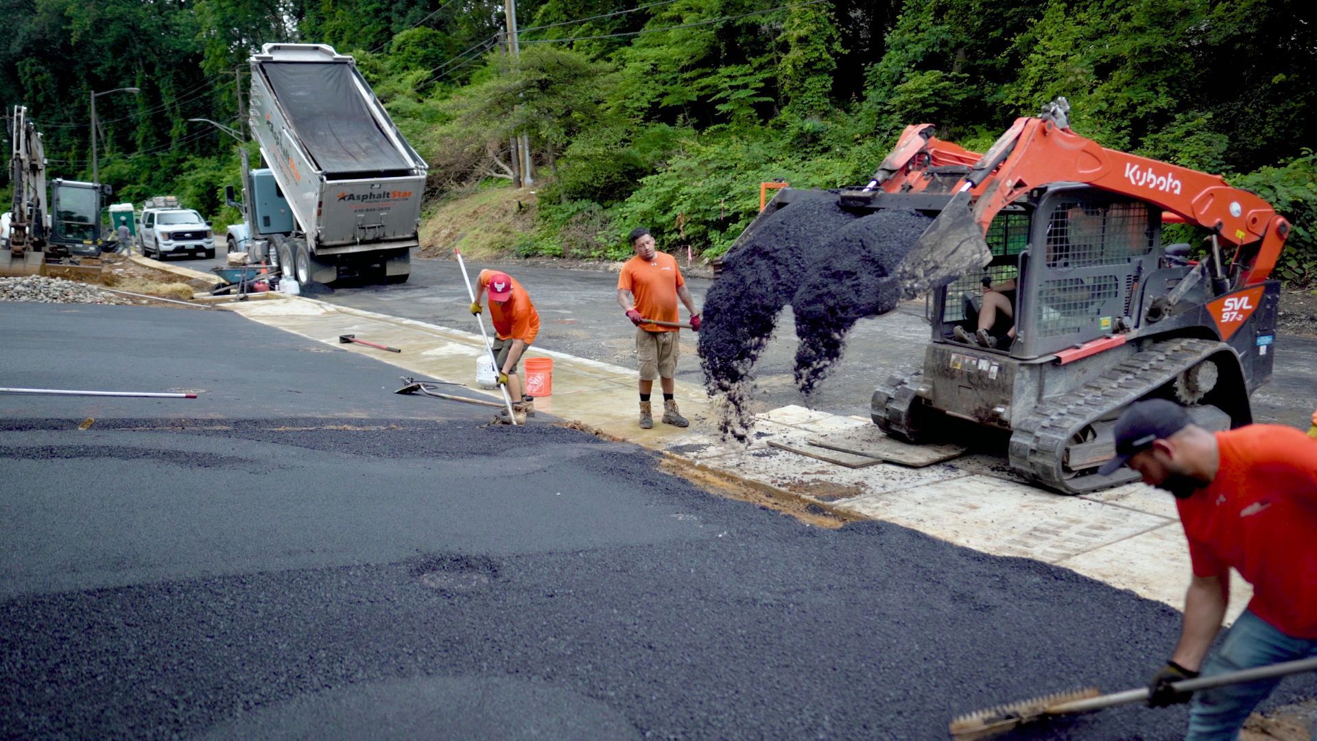 Workers lay fresh asphalt on a road using a dump truck and a skid-steer loader at a sunny construction site.