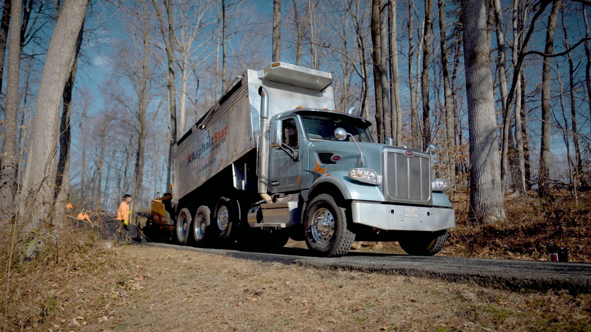 A light-colored dump truck travels along a road through a forest with workers standing nearby.