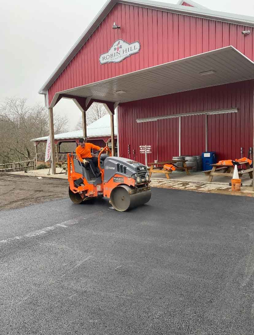 A person in bright orange clothing operates an orange asphalt roller to pave a driveway in front of a red barn building.