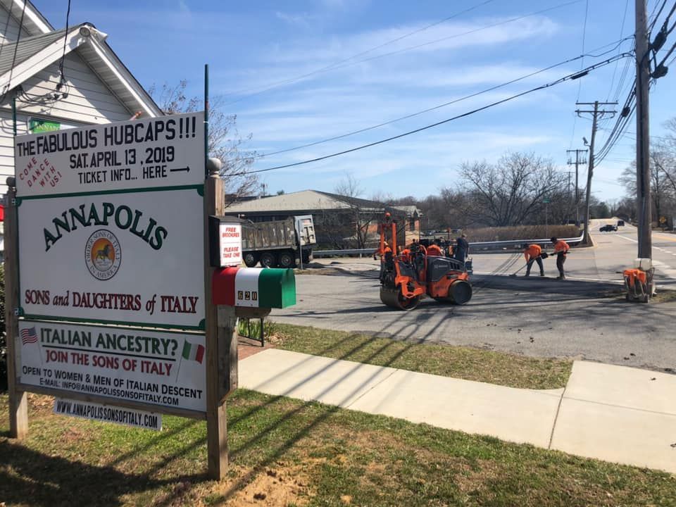 A sign for the Annapolis Sons and Daughters of Italy, with road construction workers and machinery visible in the background.