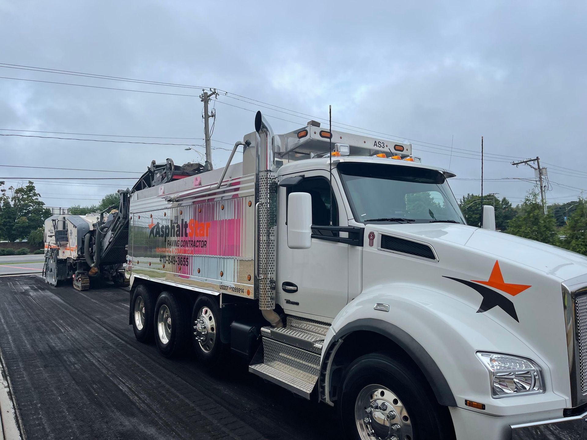 A white construction truck with an orange star logo parked on freshly paved asphalt next to a milling machine.