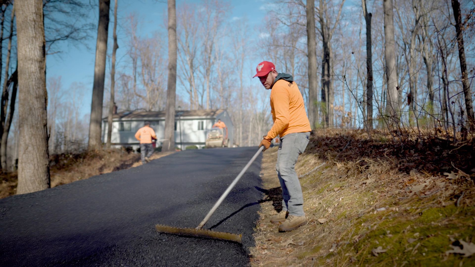 A worker in an orange sweatshirt and red cap uses a rake to smooth fresh asphalt on a driveway in a wooded area.