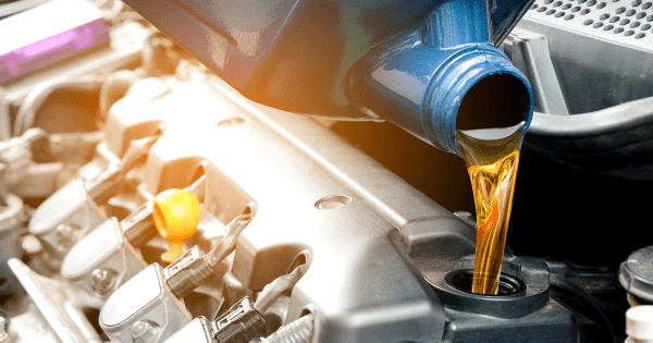 Oil being poured from a blue bottle into an engine, with a yellow cap and metal components nearby.