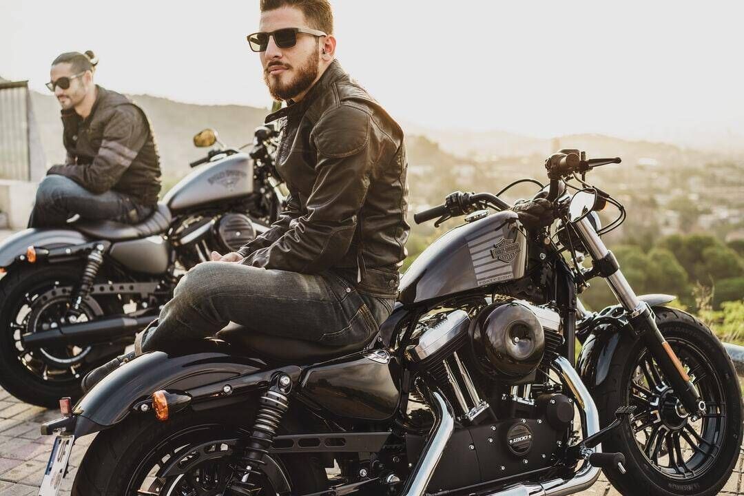 Two men on black motorcycles outdoors, one in a leather jacket and sunglasses in the foreground