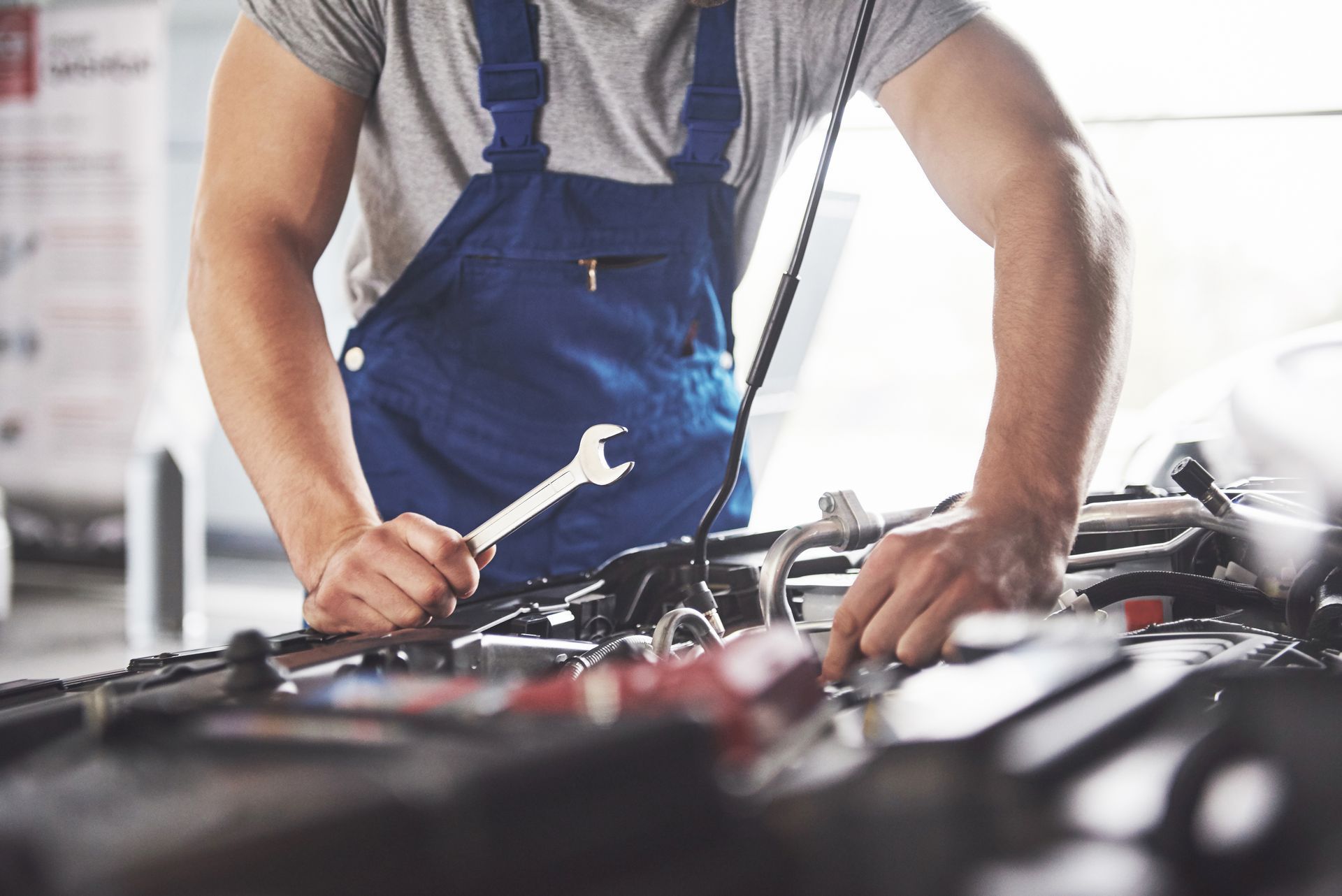 Mechanic in blue overalls working under a car hood with tools in a garage