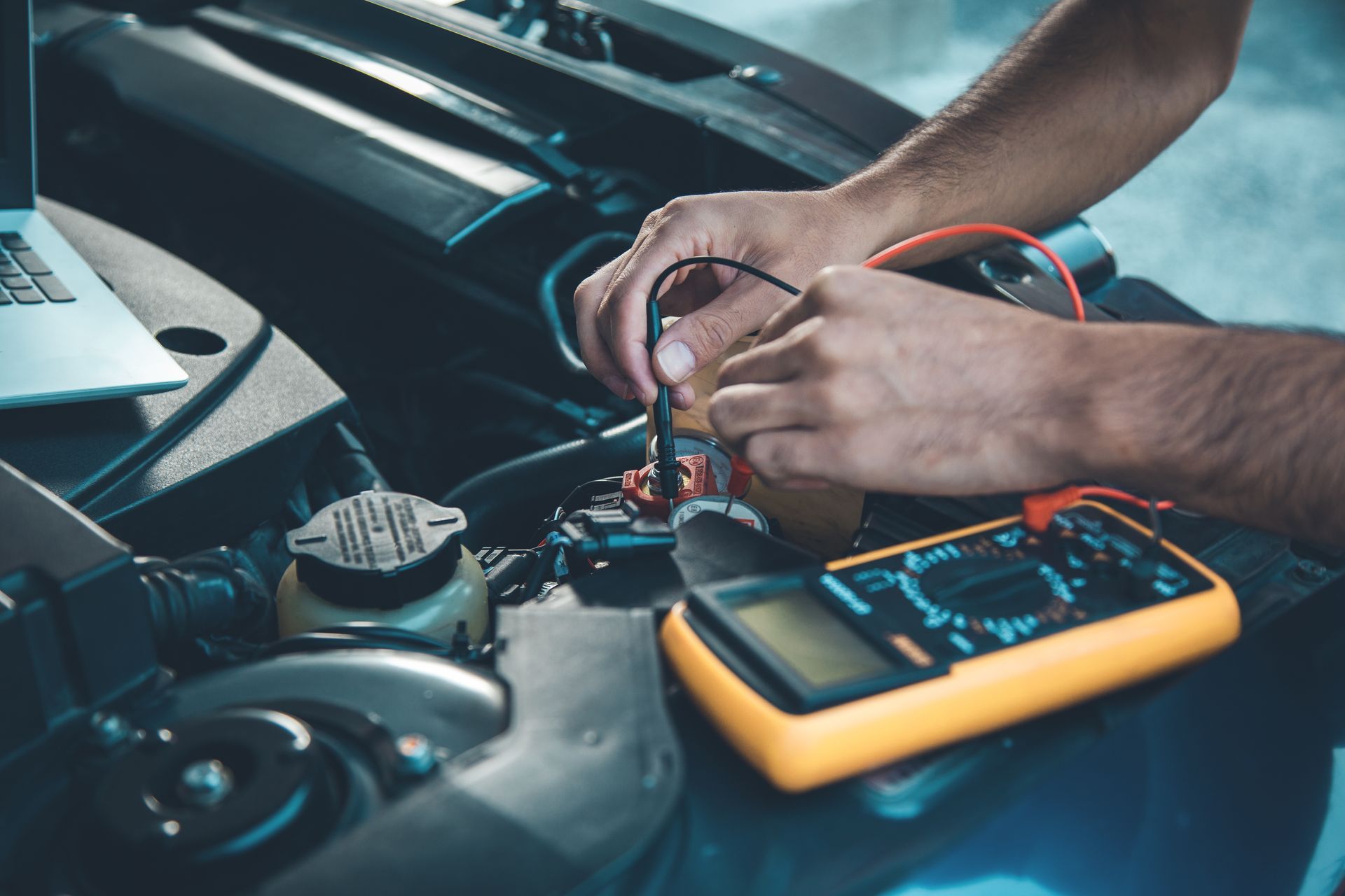 Mechanic’s hands testing wires in a car engine bay with a yellow multimeter