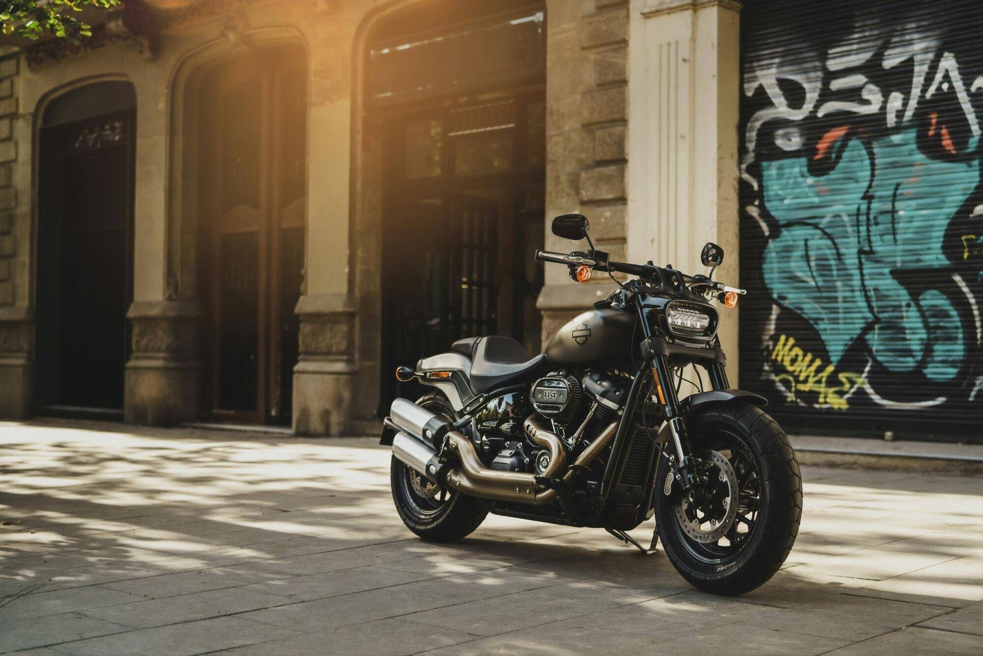 Black motorcycle parked on a sunlit city street beside a graffiti-covered wall