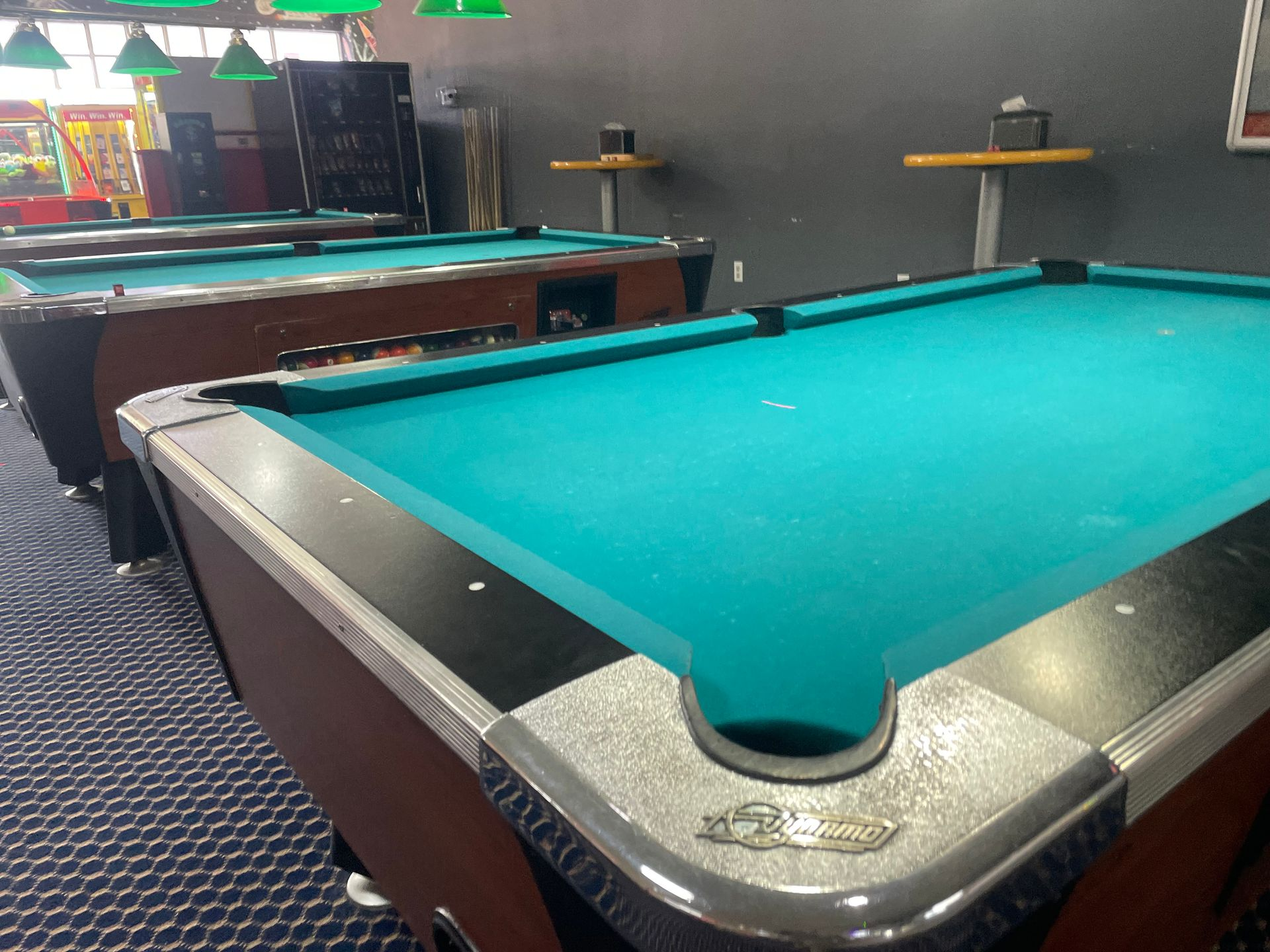 Pool tables in a dimly lit room, with green felt and dark brown frames, along a wall.