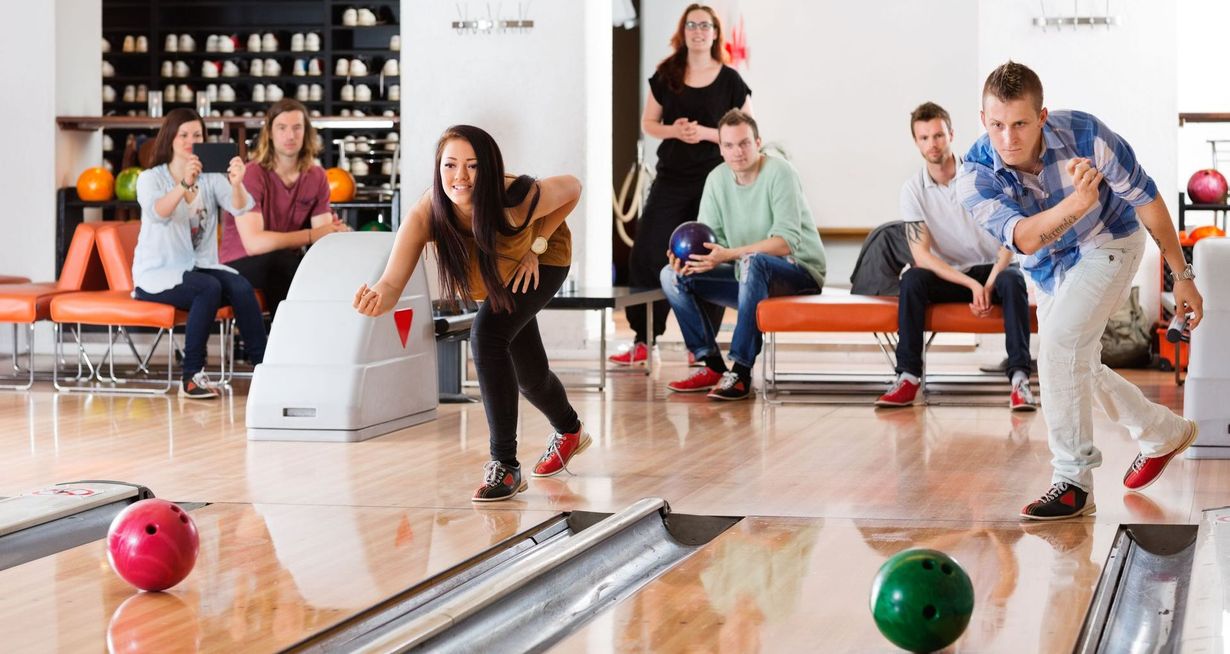People bowling in a brightly lit bowling alley. A woman throws the ball as others watch and prepare to bowl.