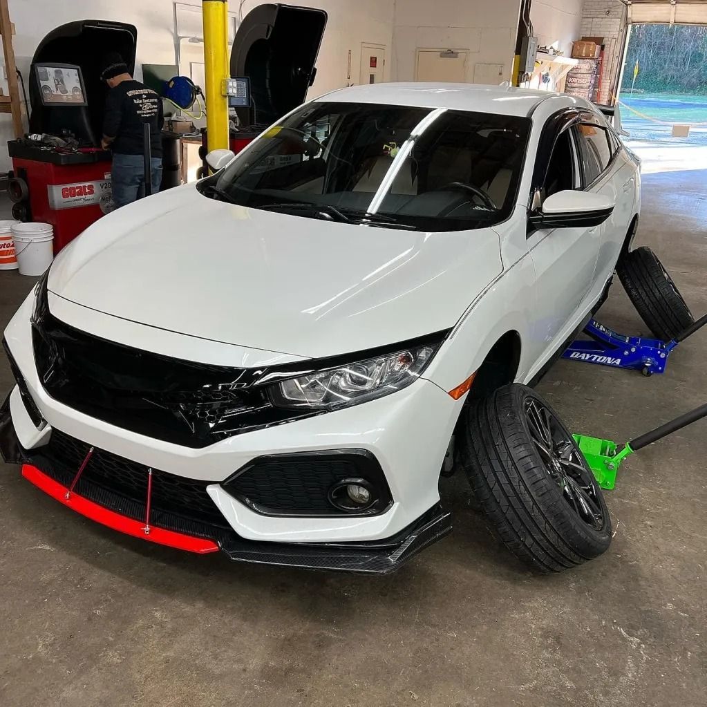 White car on a garage lift with front bumper removed and wheels turned outward, undergoing repair
