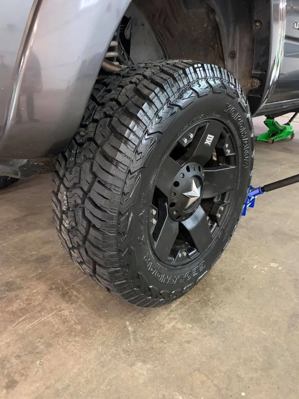 Close-up of a black off-road truck tire and wheel on a garage floor, with the vehicle lifted.