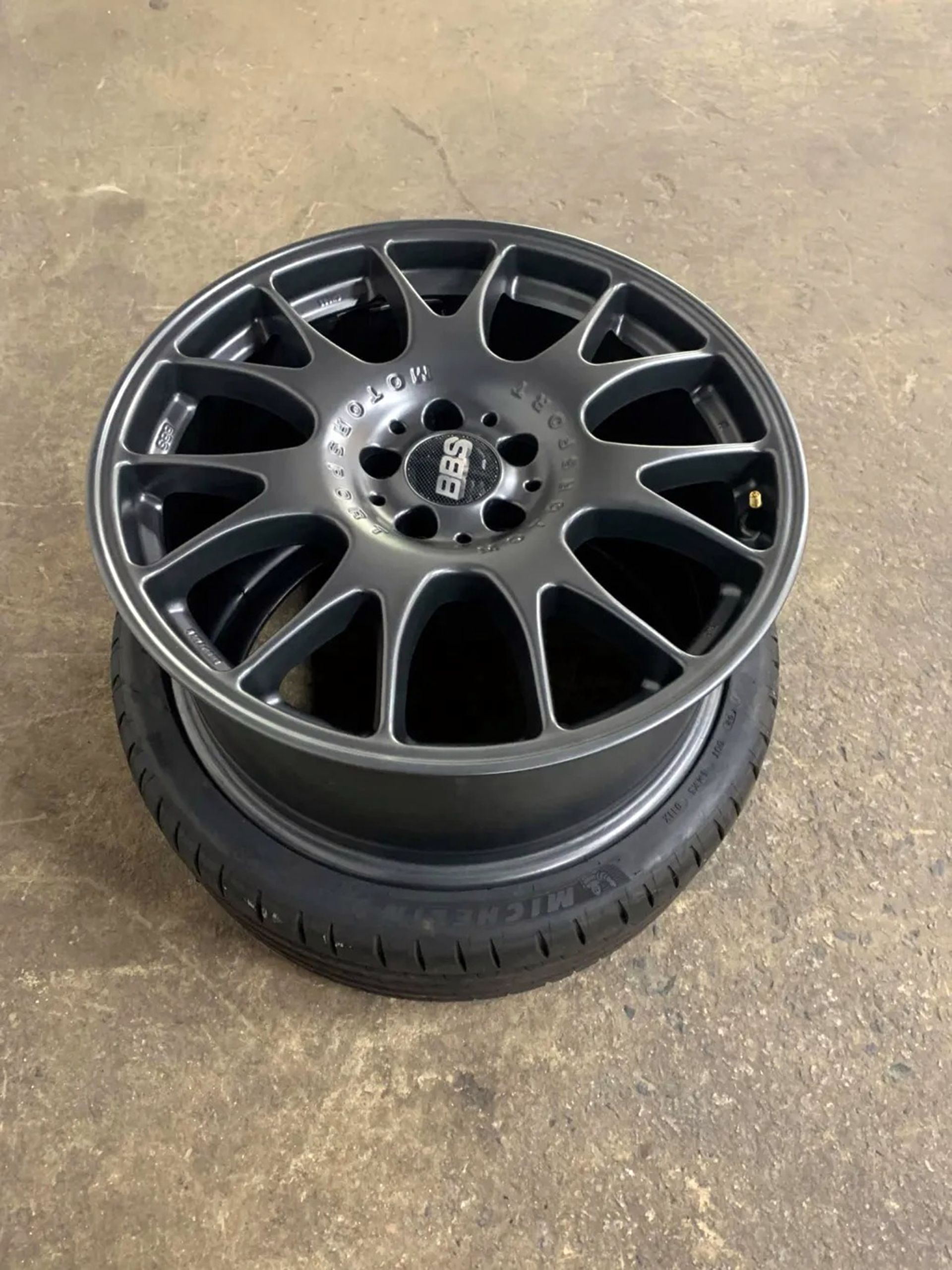 Black alloy car wheel rim with tire, lying on a concrete floor.
