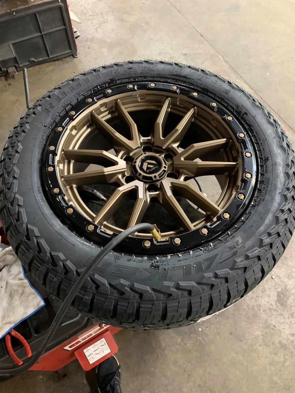 Off-road tire mounted on a bronze alloy wheel, resting on a red floor jack in a garage.