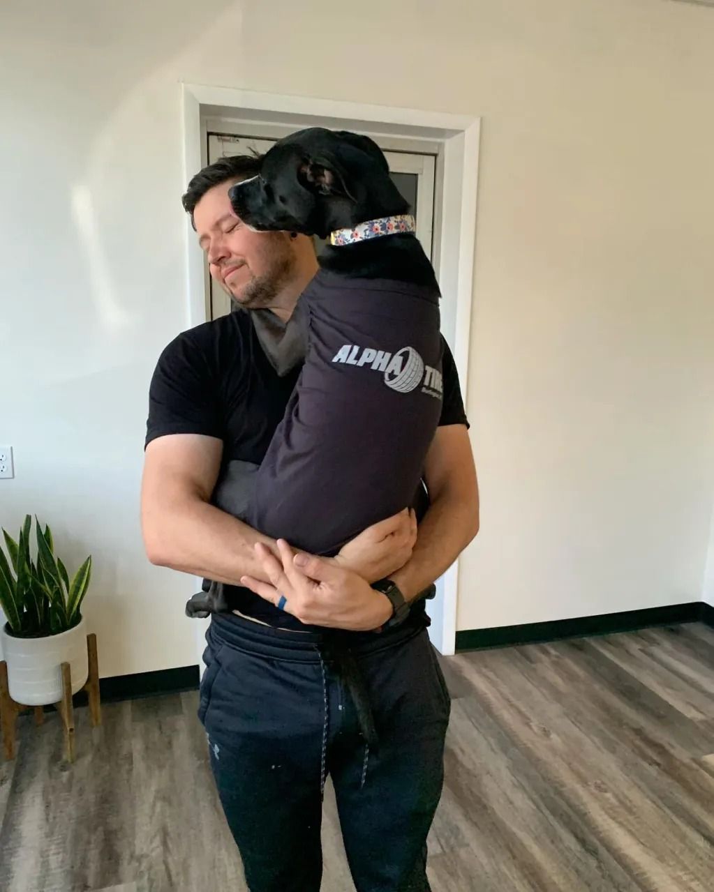 Man holding a black dog in a bright room with wood floors, both wearing dark shirts.
