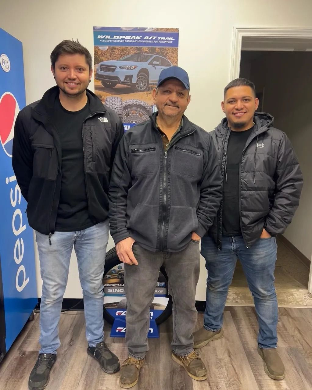 Three men in jackets standing indoors beside a Pepsi banner and a vehicle poster.