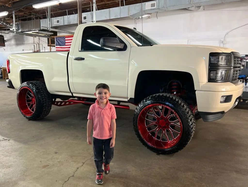 Young child standing in front of a lifted white pickup truck with red rims in a garage
