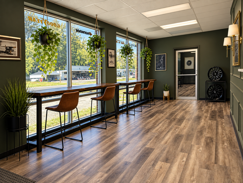 Modern office lounge with wood floors, large windows, plants, and chairs along a high counter.