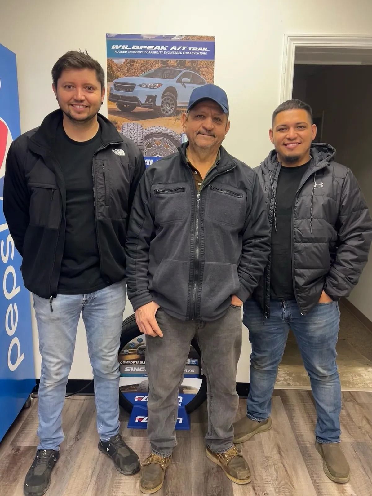 Three men in dark jackets stand indoors in front of a poster and blue banner.