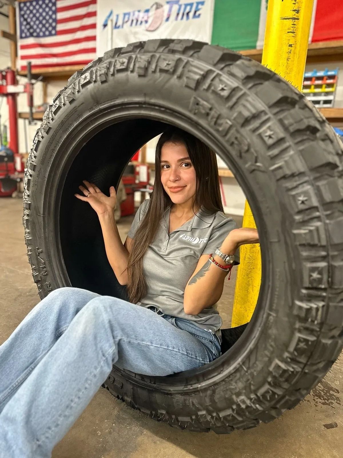 Woman sitting inside a large off-road tire in a tire shop, with an American flag in the background.