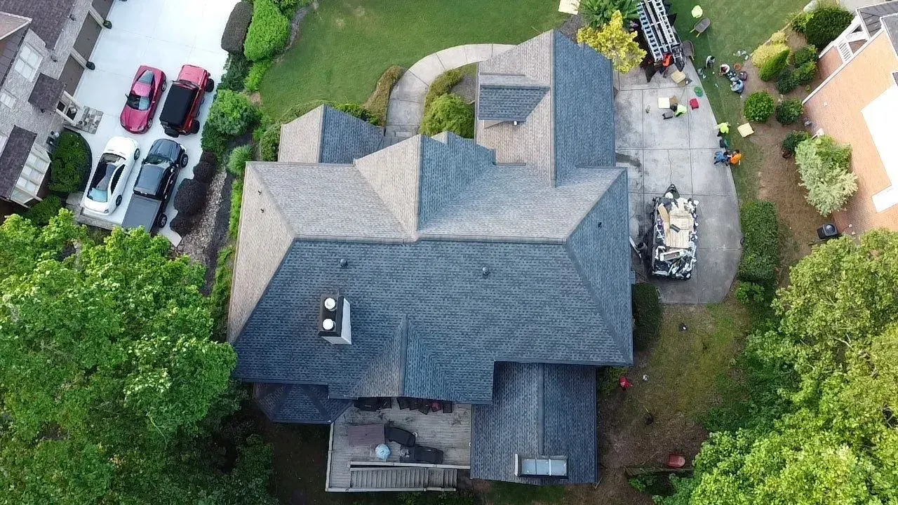 Aerial view of a house with a multi-tone roof, cars in the driveway, and surrounded by trees and lawn.