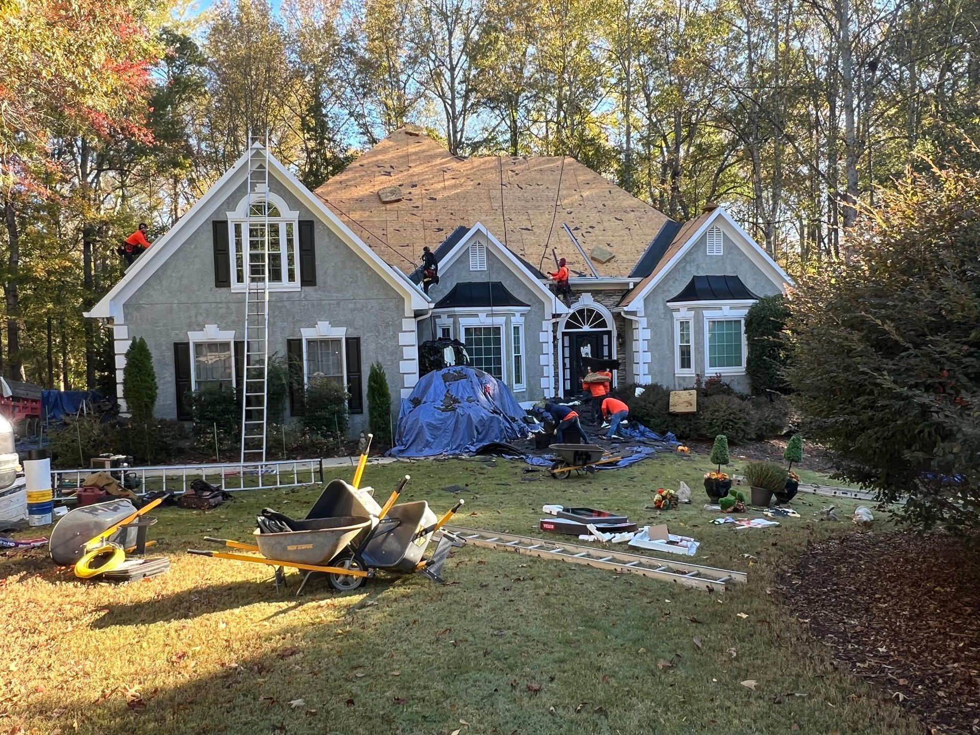 House with roof being repaired, workers, ladders, tarps, tools, and debris on lawn.