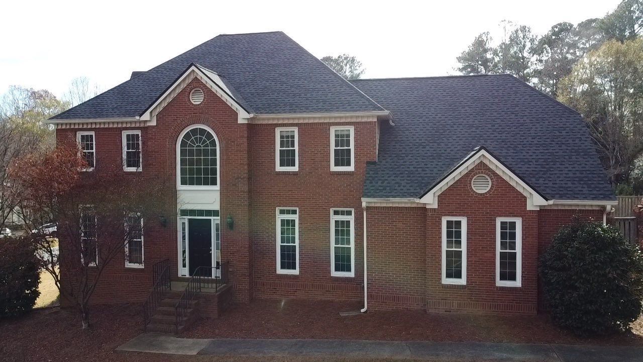 Two-story brick house with dark gray roof and multiple white-framed windows; front yard, sunny day.