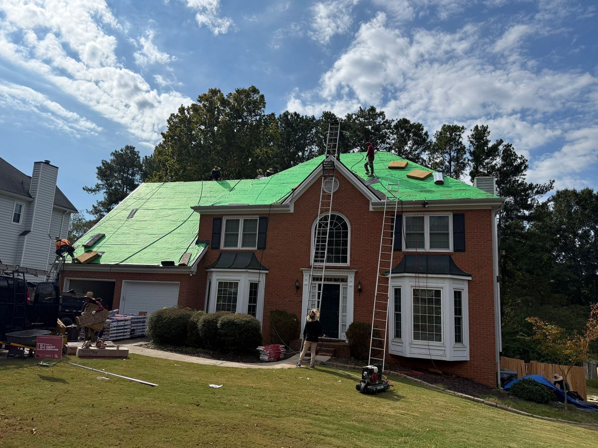 Roof being replaced on a two-story brick house. Workers on roof, green underlayment visible, ladders, and materials on the ground.