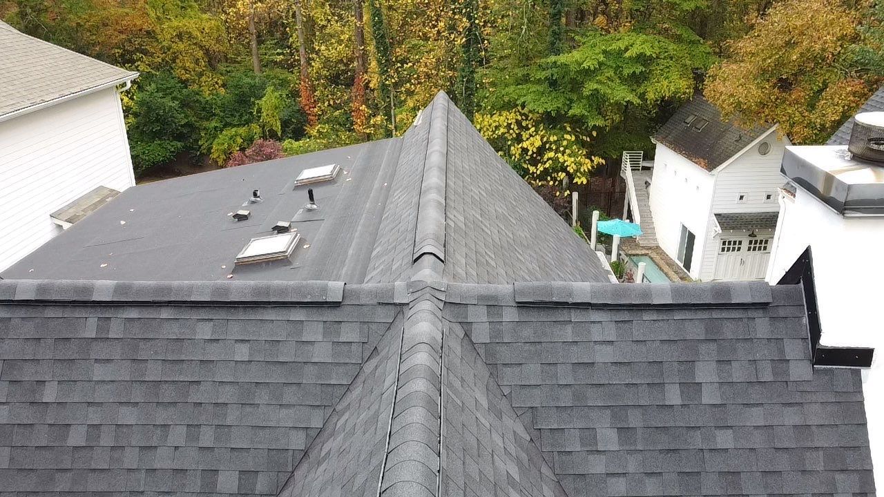 Overhead view of a dark gray shingled roof with a central ridge, surrounded by trees and houses.
