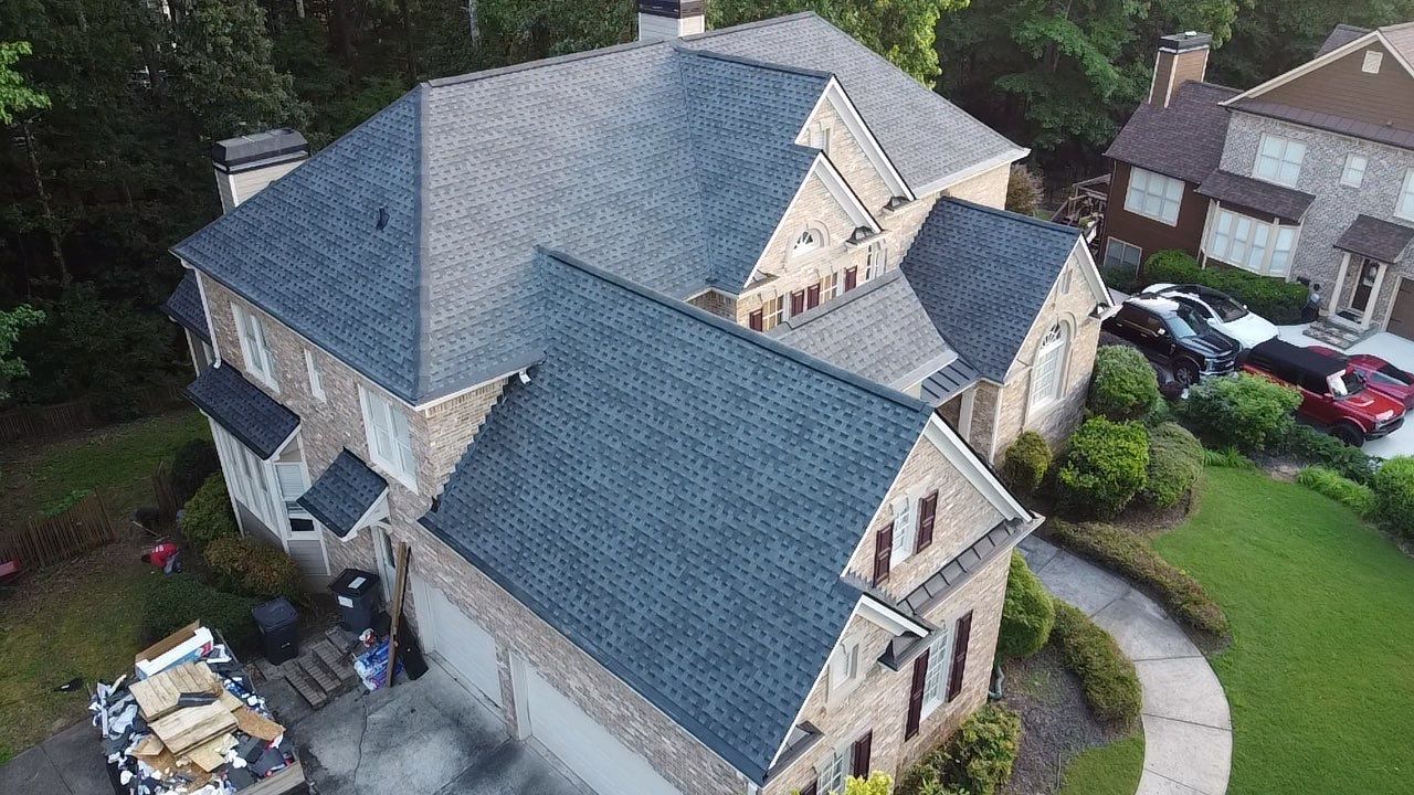 Aerial view of a two-story brick house with a dark gray shingled roof, trees and grass surrounding.